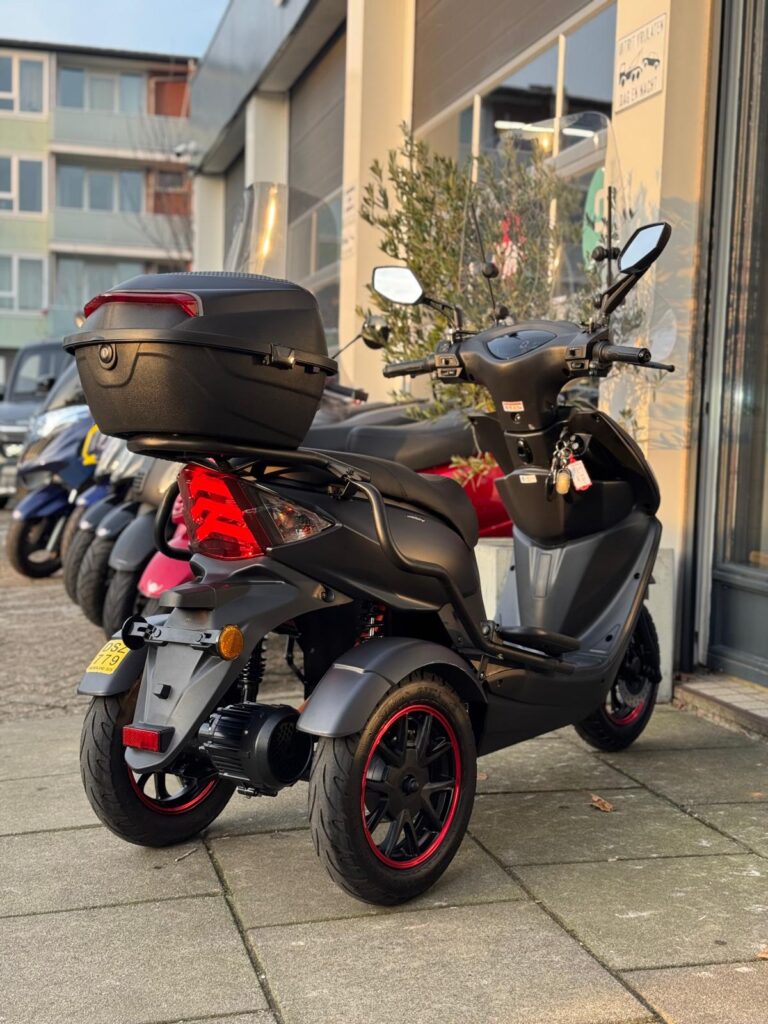 Grijze driewielscooter met topkoffer en rode velgranden, geparkeerd voor een scooterspeciaalzaak.