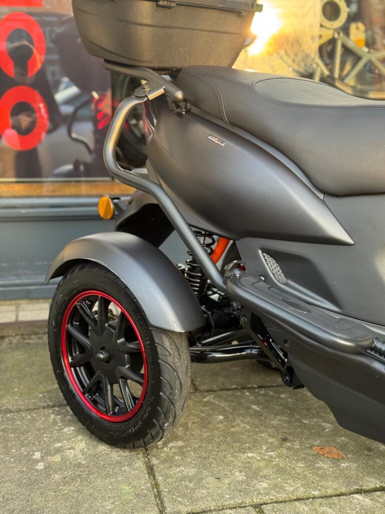 Close-up van het achterwiel en de zijkant van een matte zwarte scooter met rode velgrand en topkoffer.