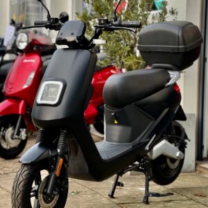 Mat zwarte elektrische scooter met topkoffer en vierkante LED-koplamp, geparkeerd bij een showroom.