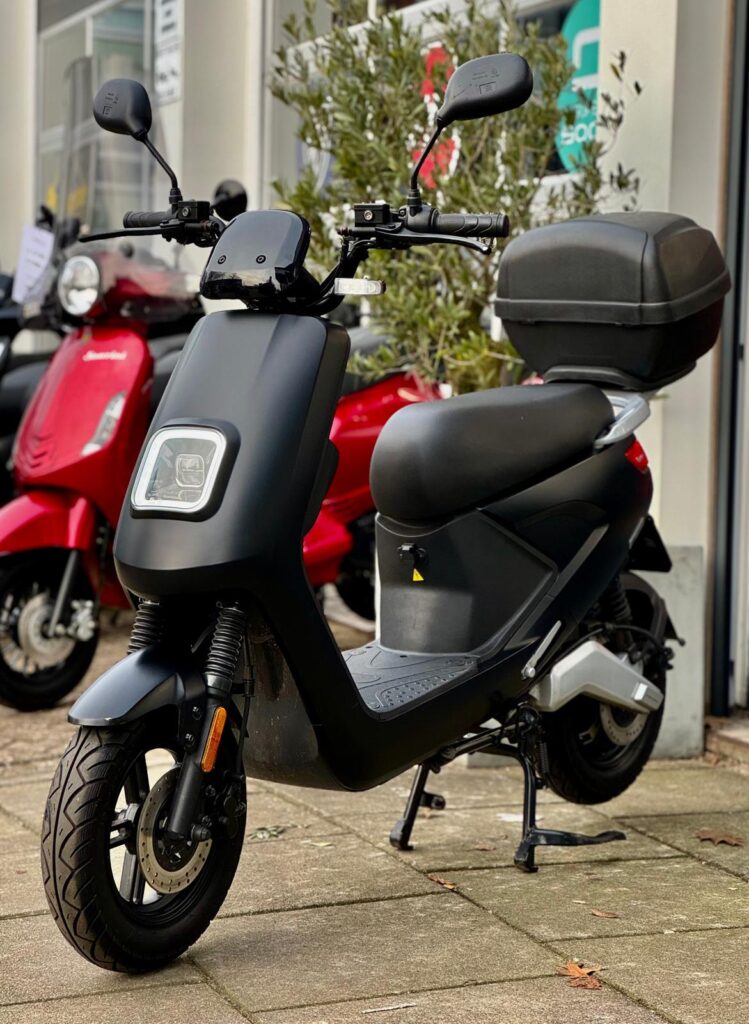 Mat zwarte elektrische scooter met topkoffer en vierkante LED-koplamp, geparkeerd bij een showroom.