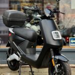 Zwarte elektrische scooter met topkoffer, geparkeerd voor een winkelraam.