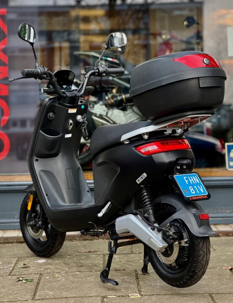 Zwarte elektrische scooter met topkoffer en blauw kenteken, geparkeerd voor een winkel.