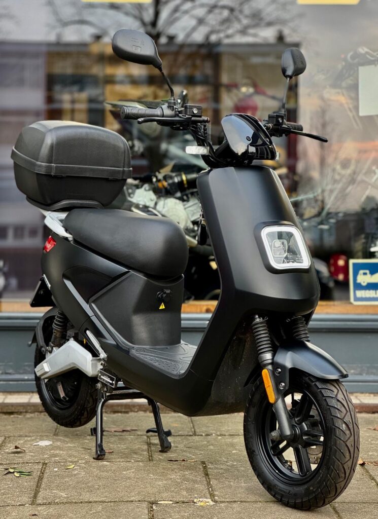 Zwarte elektrische scooter met topkoffer, geparkeerd voor een winkelraam.