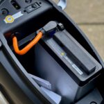 Onder zadel van e-scooter met verwijderbare accu en oranje laadsnoer aangesloten.