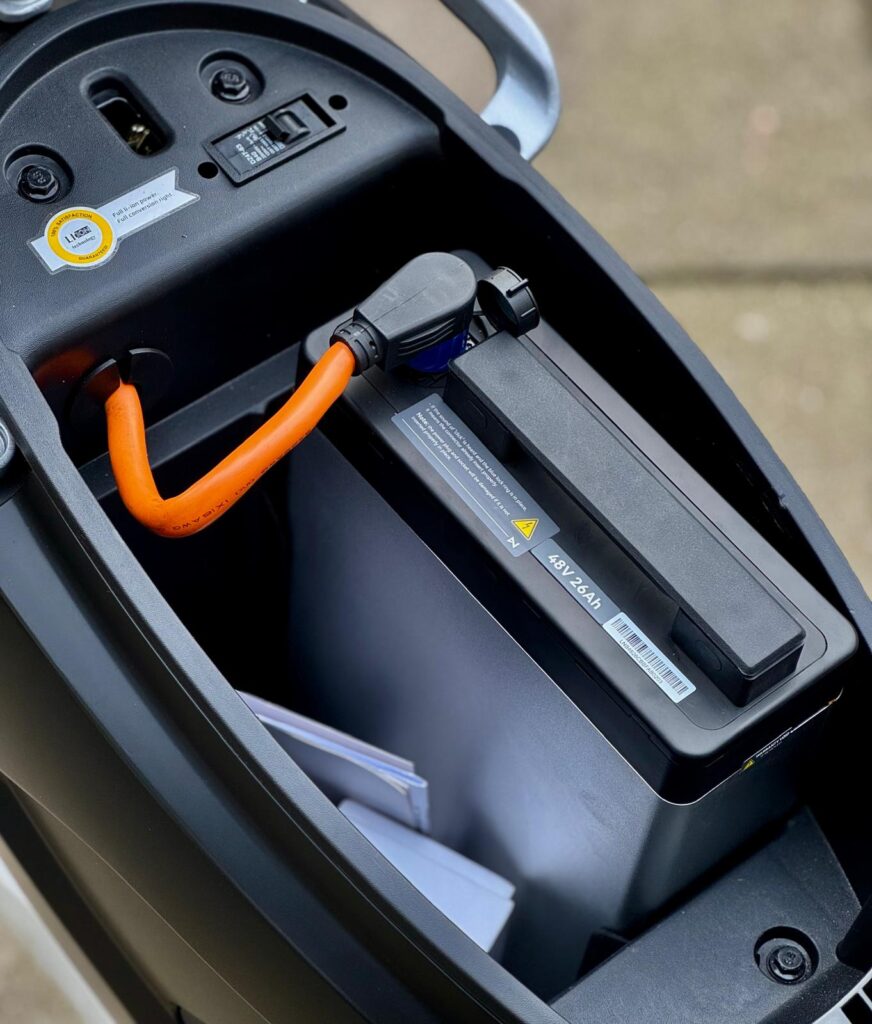 Onder zadel van e-scooter met verwijderbare accu en oranje laadsnoer aangesloten.