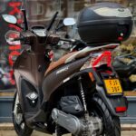 Bruine Kymco People S-scooter met ruit en topkoffer, geparkeerd voor een winkelraam op straat.