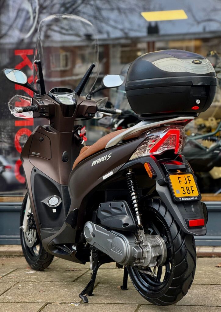 Bruine Kymco People S-scooter met ruit en topkoffer, geparkeerd voor een winkelraam op straat.
