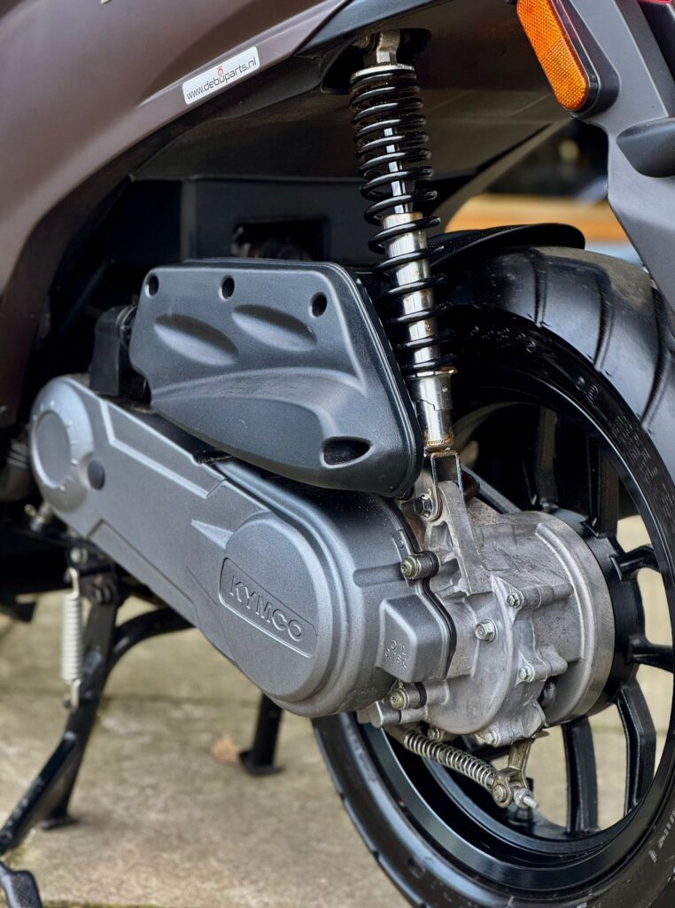 Close-up van achterwiel, schokdemper en variokap van een Kymco-scooter.