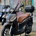 Scooter met bruin zadel, hoog windscherm en topkoffer, geparkeerd bij een scooterzaak.