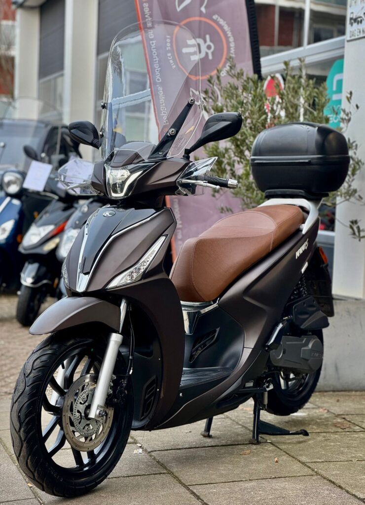 Scooter met bruin zadel, hoog windscherm en topkoffer, geparkeerd bij een scooterzaak.
