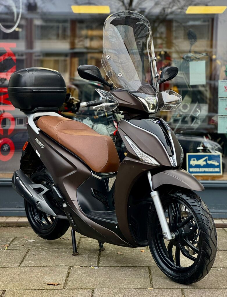 Bruine scooter met hoog windscherm en topkoffer, geparkeerd voor een winkel.