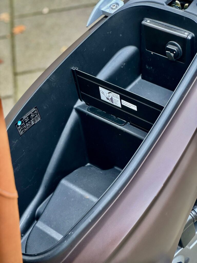 Lege opbergruimte onder het zadel van een scooter, met uitklapbare vakverdeler en waarschuwingsstickers.