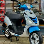 Blauw-grijze scooter met spiegels en grote koplamp, geparkeerd op de stoep voor een winkelraam.