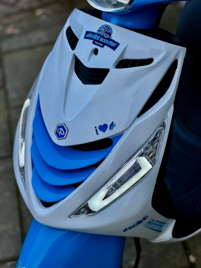 Close-up van de voorkant van een wit-blauwe scooter met LED-koplampen en Polini-stickers.