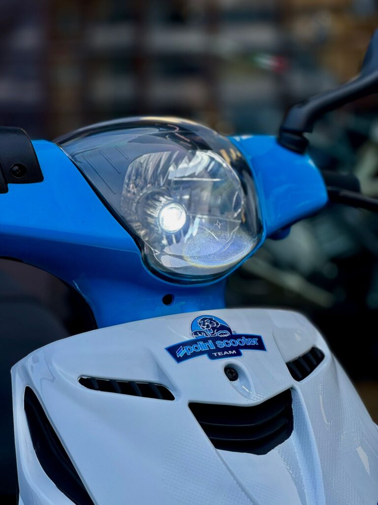 Close-up van de voorkant van een blauw-witte scooter met brandende koplamp en Polini-logo.