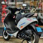 Wit-grijze scooter met blauwe accenten, op de standaard geparkeerd voor een glazen winkelpui.