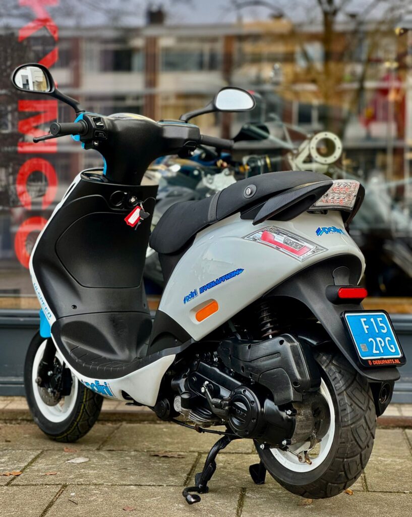Wit-grijze scooter met blauwe accenten, op de standaard geparkeerd voor een glazen winkelpui.