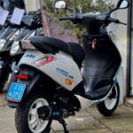 Zilverkleurige scooter met Polini-uitlaat en blauwe kentekenplaat, geparkeerd voor een scooterwinkel.