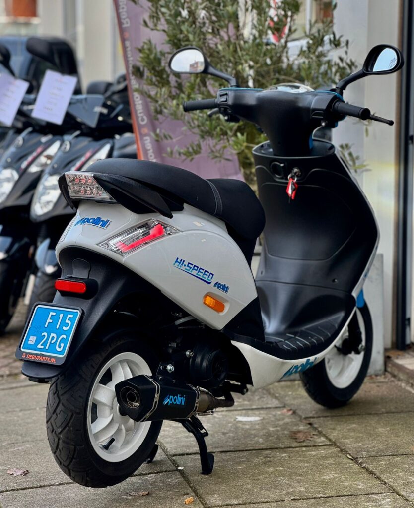 Zilverkleurige scooter met Polini-uitlaat en blauwe kentekenplaat, geparkeerd voor een scooterwinkel.