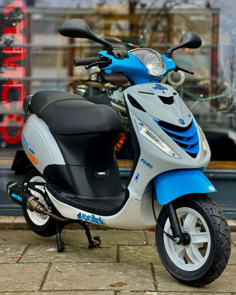 Blauw-grijze scooter met spiegels en grote koplamp, geparkeerd op de stoep voor een winkelraam.