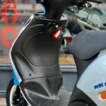 Close-up van stuur en voetenplank van zwart-witte scooter met rode sleutel in contactslot, geparkeerd voor een etalage.