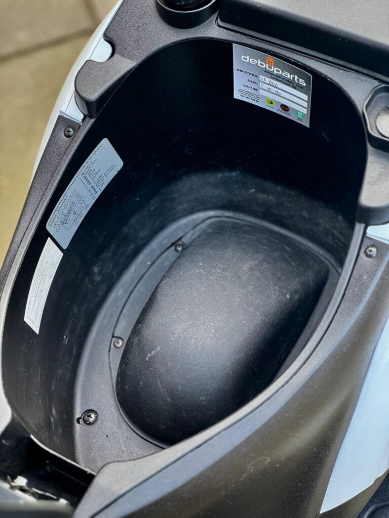 Lege onderzadel-opbergruimte van een scooter, met stickers en onderhoudslabel.