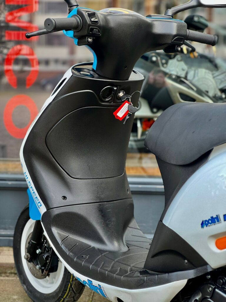 Close-up van stuur en voetenplank van zwart-witte scooter met rode sleutel in contactslot, geparkeerd voor een etalage.