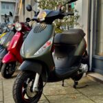Matgroene scooter met schijfrem geparkeerd voor een winkel, naast rode en gele scooters.