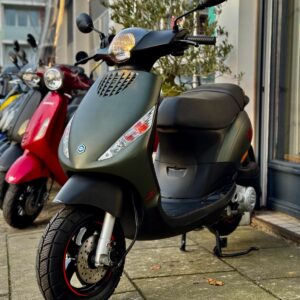 Matgroene scooter met schijfrem geparkeerd voor een winkel, naast rode en gele scooters.
