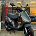 Matgroene scooter op standaard voor een winkelraam, met zonsondergang weerspiegeld in het glas.