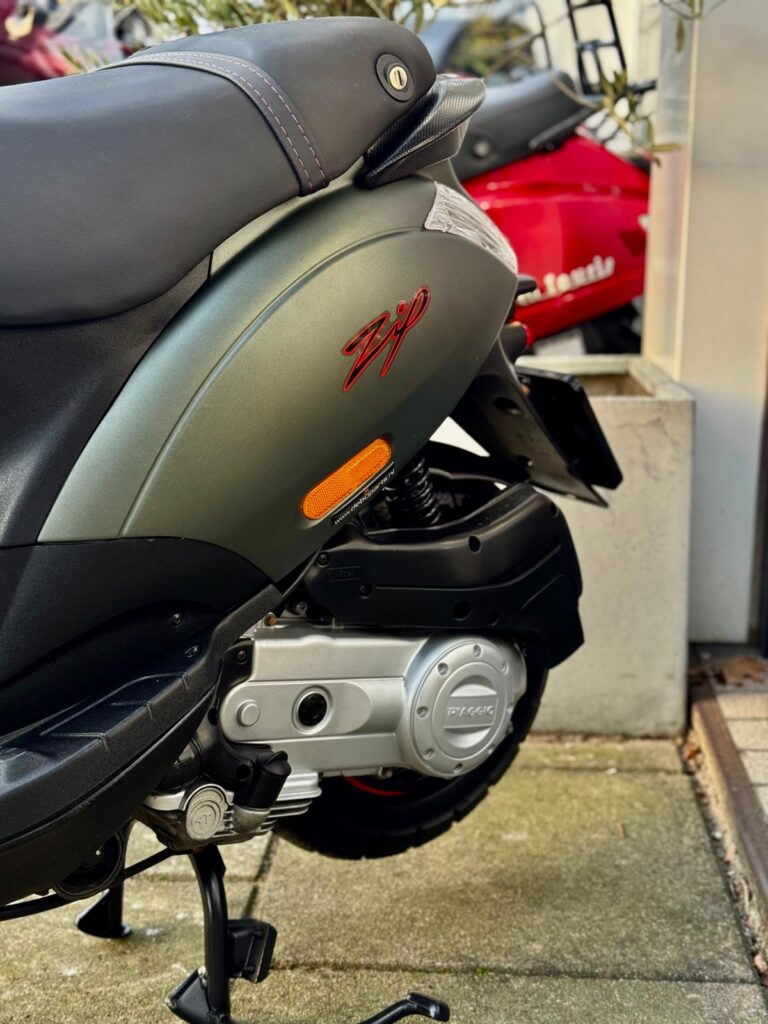 Close-up van een Piaggio Zip scooter, zijkant met zadel en motorblok, buiten geparkeerd.