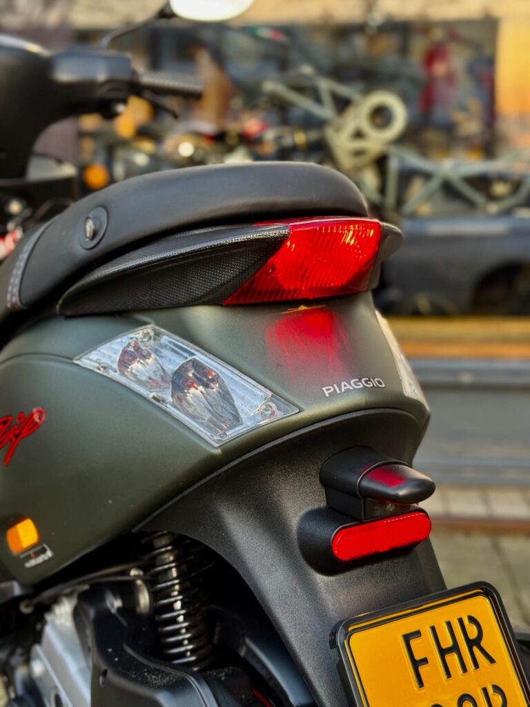 Close-up van de achterzijde van een Piaggio-scooter met rood achterlicht, knipperlichten en gele kentekenplaat.