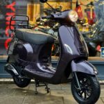 Donkerpaarse retro scooter met brandende koplamp, geparkeerd op een stoep voor een showroom.