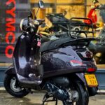 Paarszwarte retro scooter met bagagedrager en geel kenteken, geparkeerd voor een motorenshowroom.