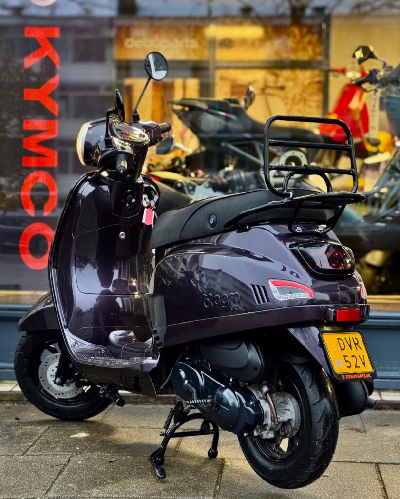 Paarszwarte retro scooter met bagagedrager en geel kenteken, geparkeerd voor een motorenshowroom.