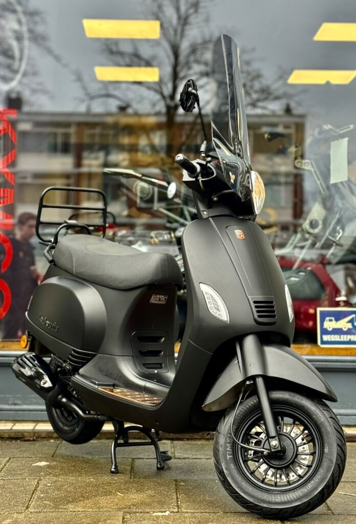 Matzwarte scooter met hoog windscherm, geparkeerd voor een winkelruit op straat.