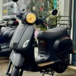 Matzwarte scooter met windscherm en brandende koplamp, geparkeerd voor een winkel op nat trottoir.