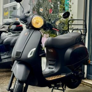 Matzwarte scooter met windscherm en brandende koplamp, geparkeerd voor een winkel op nat trottoir.