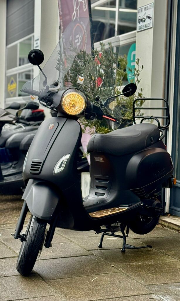 Matzwarte scooter met windscherm en brandende koplamp, geparkeerd voor een winkel op nat trottoir.