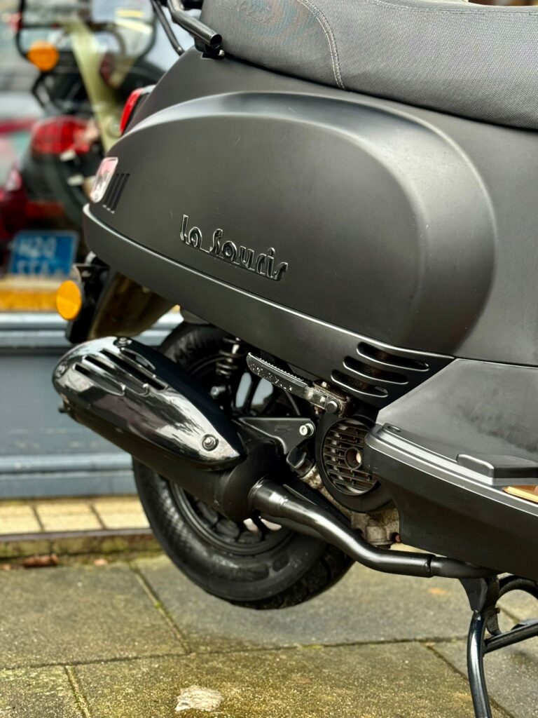 Close-up van de achterzijde van een matte zwarte La Souris scooter met uitlaat en wiel.