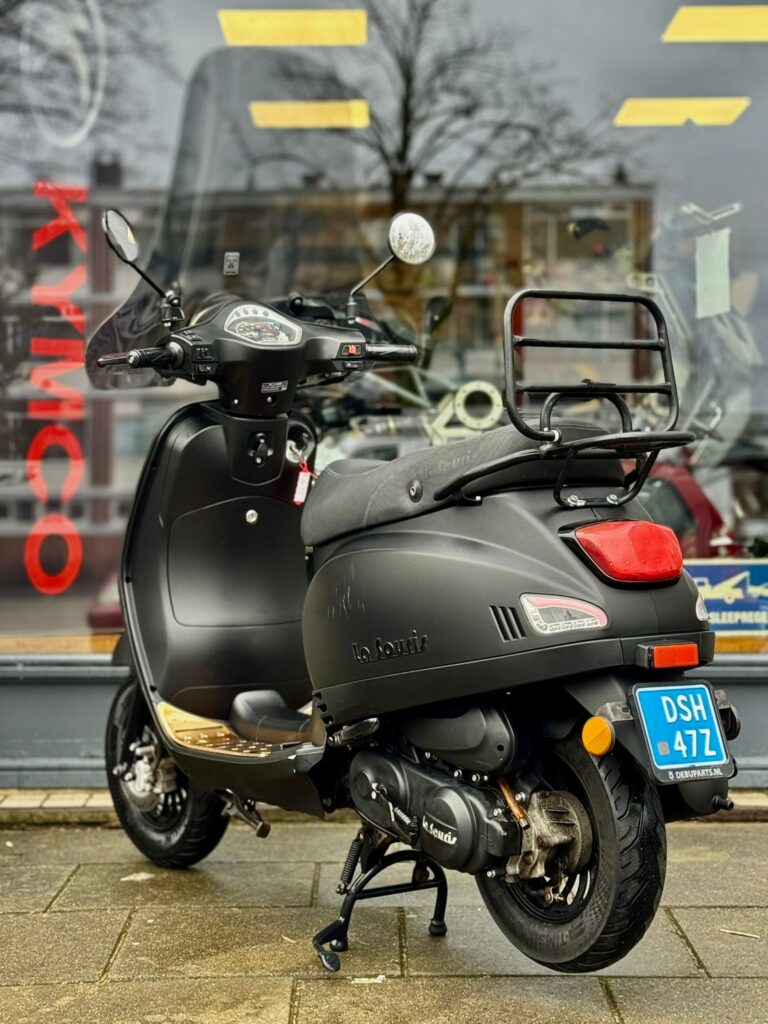 Matte zwarte scooter met windscherm en bagagedrager, geparkeerd voor een winkelraam.