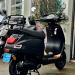 Matzwart retro-scooter met bagagedrager en blauwe kentekenplaat, geparkeerd voor een scootershop.