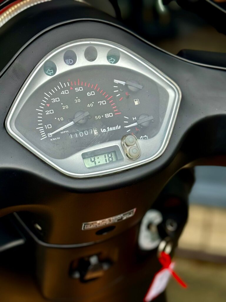 Close-up van een scooterdashboard met snelheidsmeter, brandstof- en temperatuurmeter en klein digitaal klokje.