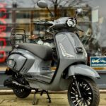 Grijze retro-stijl scooter met LED-verlichting, geparkeerd voor een etalage op de stoep.