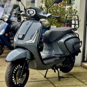 Grijze retro-look scooter met LED-koplamp en bagagedrager, geparkeerd voor een scootershop.
