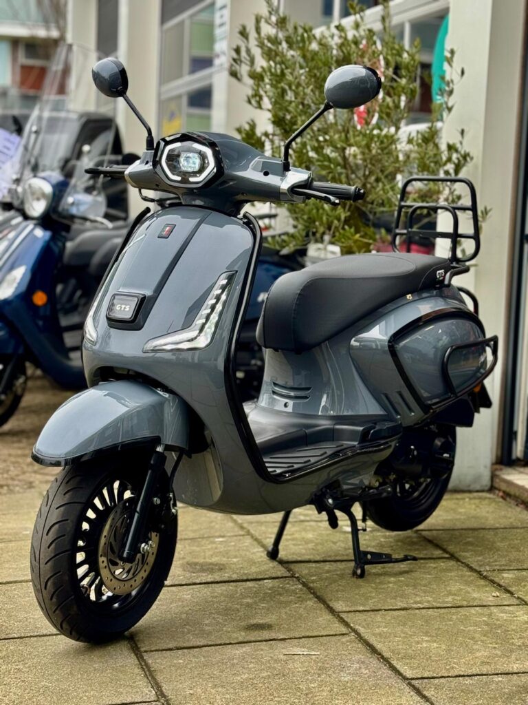 Grijze retro-look scooter met LED-koplamp en bagagedrager, geparkeerd voor een scootershop.