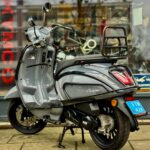 Grijze retro-scooter met achterdrager geparkeerd voor een winkelraam, zijaanzicht op het trottoir.