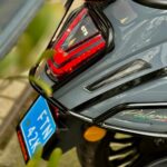 Close-up van de achterkant van een grijze scooter met rood achterlicht, bagagerek en blauwe kentekenplaat.