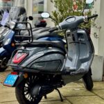 Grijze retro scooter met blauwe kentekenplaat geparkeerd voor een winkel, gezien schuin van achteren.