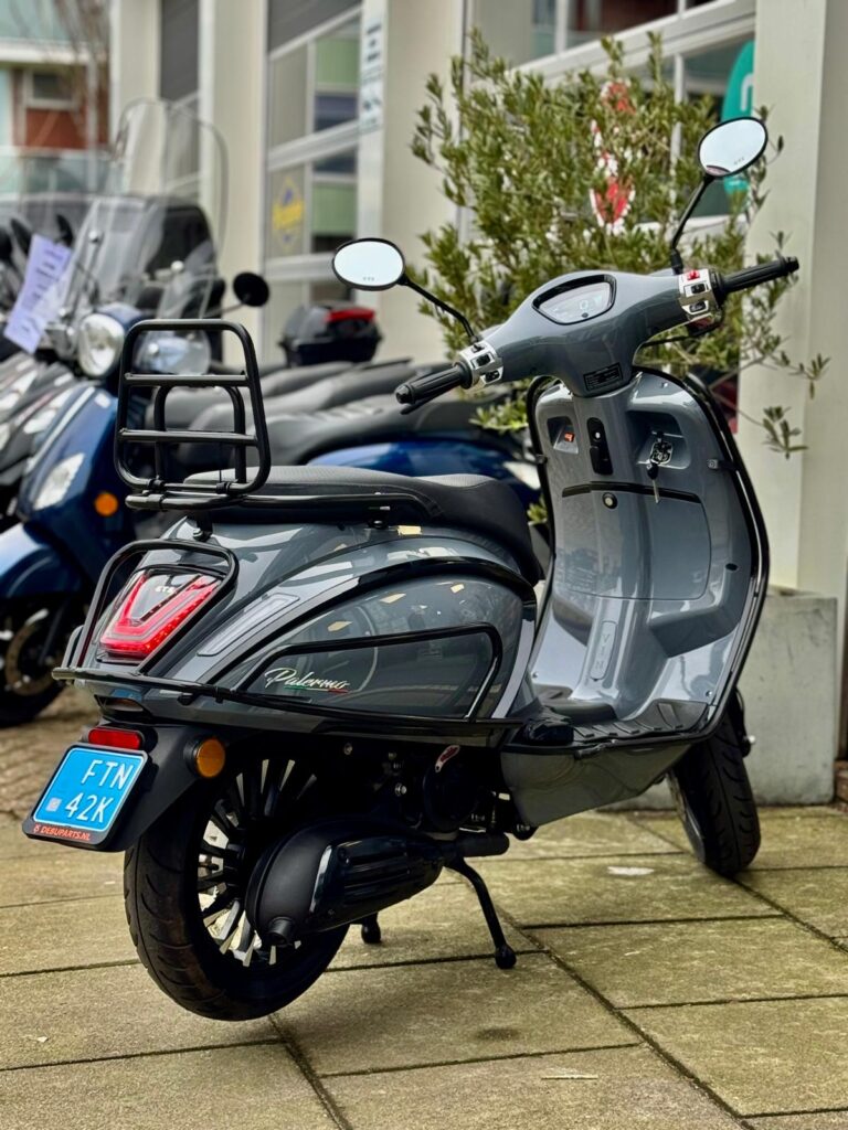 Grijze retro scooter met blauwe kentekenplaat geparkeerd voor een winkel, gezien schuin van achteren.